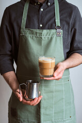 Barista makes coffee. A man holds a milk jug and a glass of coffee. The guy makes cappuccino, pouring whipped milk into espresso. Barista in a kitchen apron. Equipment for professional service