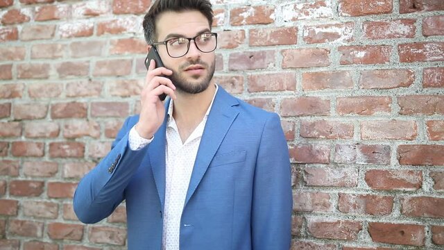 happy businessman standing agains a brick wall, answering his phone, smiling and looking around