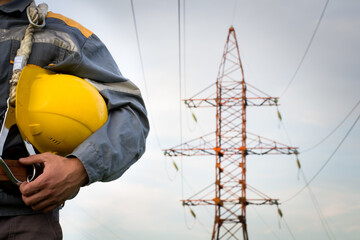A young electrician is holding a protective helmet. Power plant worker on the background of an electric pylon. Electric support 110 kilovolts. Emergency service for repairing electrical networks. 