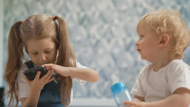 little children of different ages, show each other tongue, girl with pigtails holds smartphone, boy is blond - bottle with dummy, they sit on floor in living room, brother and sister smiling close up