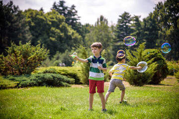 Fototapeta premium Boy blowing soap bubbles while an excited kid enjoys the bubbles. Happy teenage boy and his brother in a park enjoying making soap bubbles. Happy childhood friendship concept