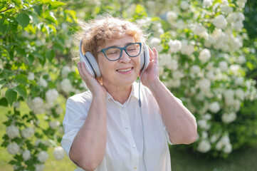 Happy old woman walks in the park of blooming apple trees and listens to music on headphones. A female pensioner is smiling and enjoying a warm summer day in the garden. Advanced older generation.