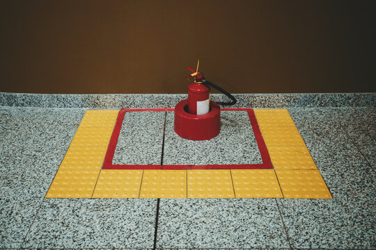 A Red Powder Fire Extinguisher Indoors Of An Airport Or A Mall In A Special Yellow Zone Made Of Tactile Tiles On The Marble Floor Near A Brown Wall