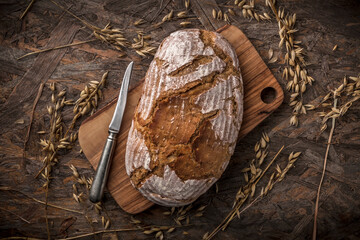 Fototapeta na wymiar Wholegrain rustic fresh bread with knife on cutting board arrangement with rye and wheat stalks on old rustic wooden table studio shot