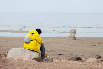 Obraz premium Burdened by unseen worries. Sad lonely man in yellow raincoat sits on stone at seashore looking forward.