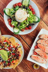 Vertical photo of a goat cheese salad, nachos with guacamole and meat and bread with tomato on a wooden table