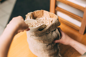 Woman measuring brown rice dosage, taking one plastic scoop of this healthy cereal out of jute clouth bag. Shopping in bulk.