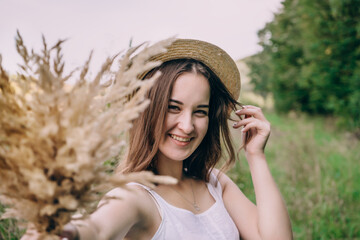 Happy young woman in a white dress in a straw hat. Smiling girl with a bouquet of dried flowers. Girl walks in a green park on a spring day.