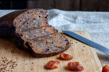 Black rye homemade bread in the form of a loaf with the addition of dried apricots, prunes, nuts on a wooden board decorated with flax seeds, dried apricots and a knife on a wooden board on canvas.
