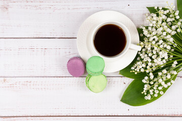 Obraz na płótnie Canvas Colorful macaroons, a cup of coffee and lilies of the valley on wooden background, close-up flat lay. Concept of breakfast and spring time. Copy space.