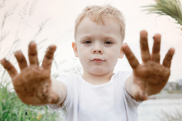 A boy shows his dirty hands after playing with sand. On the hands of the remnants of wet sand. a boy in a white T-shirt