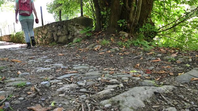 Imagine close to the ground of a path: you can see the boots of a woman who is walking with a red backpack on her shoulders.