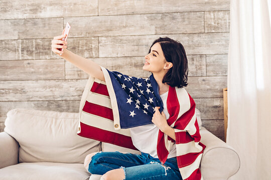 Lifestyle portrait of happy young woman taking selfie with American Flag by phone on July 4th, Independence Day - Powered by Adobe