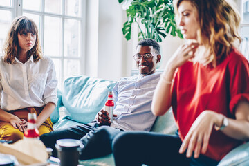 Cheerful black man drinking holding bottle in hand sitting on sofa
