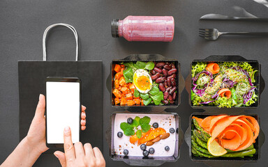 Female hand holding phone mock up white screen over vegan healthy food package vegetable delivery boxes black table background. Veggie diet nutrition menu order plan mobile app ad. Flat lay top view