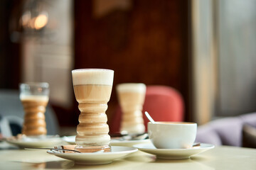 Latte glass with layered latte, cappuccino or mocha with foam on table in cafe with milk saucer and spoon. Three cups of coffee. Food and drink toning image.