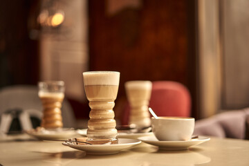 Latte glass with layered latte, cappuccino or mocha with foam on table in cafe with milk saucer and spoon. Three cups of coffee. Food and drink toning image.