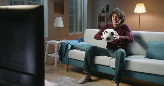 Expressive south asian football team supporter emotionally watching a soccer game. Hipster guy with curly hair watching tv at home alone 4k footage