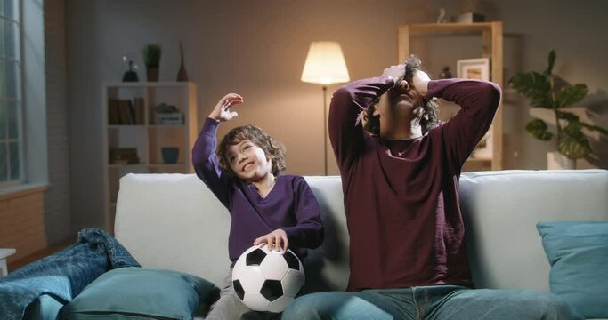 Authentic young cute asian brothers with curky hair watching soccer together, father and son reacting to vicories or defeats of their team, expressing emotions - family time concept 4k footage