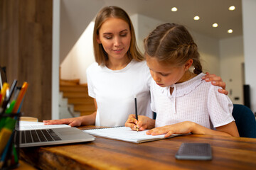 Fototapeta premium Joyful mother helping cute daughter to study at home