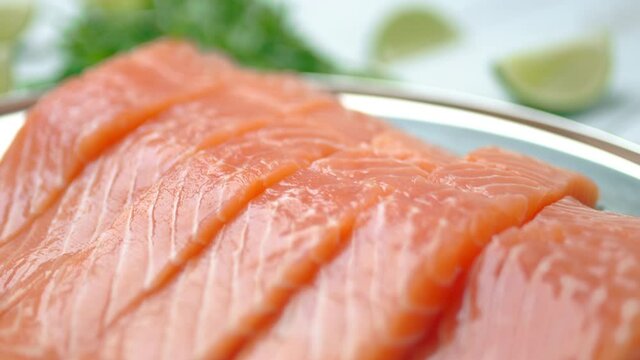 Close-up slow-motion of sliced fresh raw red salmon.
Raw salmon in a silver plate on the wooden table with lemon and herbs/greens ingredients on the side. Concept of healthy seafood. Salmon in 4K.
