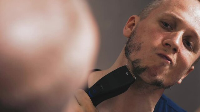 slow motion closeup man winces at himself in mirror and begins to shave off for gray and black beard electric hair machine after removing self-isolation and quarantine regime, hair flying to sides