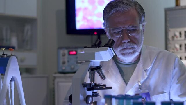 A oncology research scientist in a laboratory ponders the work he is doing with a display of cancer cells on the monitor in the background.