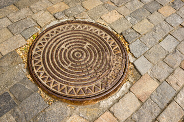 An iron manhole from a well against the background of a road made of stone paving stones. Street, road surface.