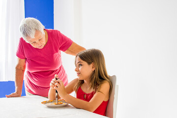 Elderly grandmother guides granddaughter feeding at table with affection