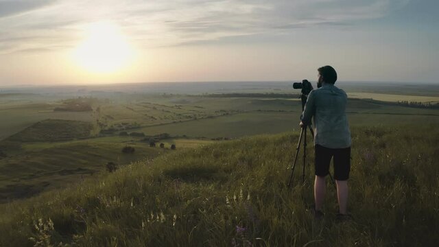 Back view of competent photographer in casual clothing standing on green hill with tripod and digital camera and doing photos of amazing sunset. Concept of adventure and nature beauty.