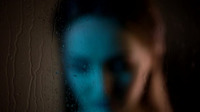 Close up shot of dreamy sad attractive caucasian woman standing near steamy window glass looking at rain drops falling