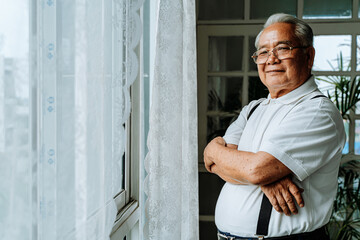 Positive plump senior Asian male smiling for camera and crossing arms while resting near window in cozy room at home - 357966202