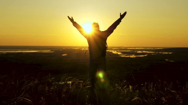 a traveler with a backpack stands on edge of mountain in rays of dawn, raises his hands in air and enjoys victory, beautiful sun and landscape. free male tourist travels alone in park