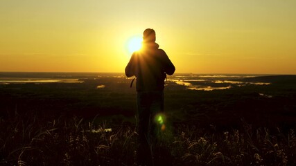 a traveler with a backpack stands on edge of mountain in rays of dawn, raises his hands in air and enjoys victory, beautiful sun and landscape. free male tourist travels alone in park