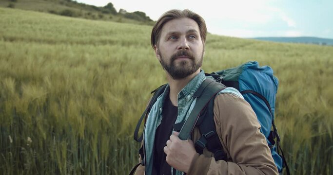 Happy bearded man with stylish haircut walking at green field with big backpack. Mature photographer enjoying beautiful nature on fresh air.