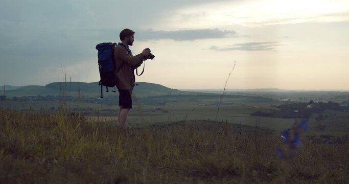 Full length portrait of travel photographer with backpack using digital camera for shooting nature while standing at green hill. Bearded man enjoying working process on fresh air.