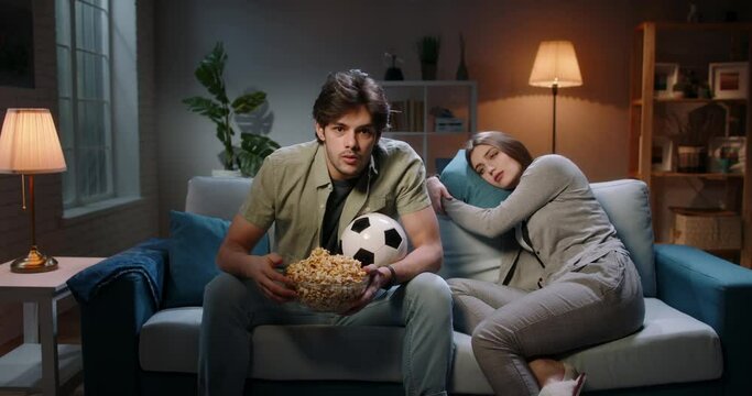 Couple watching football at home. Emotional guy watching his team play and emotionally reacting while his girlfriend is lying on couch extremely bored 4k footage