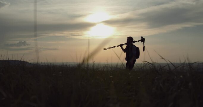 Nature photographer in silhouette carrying on backpack and tripod for taking pictures on high hill. Mature man walking outdoors during amazing sunset.
