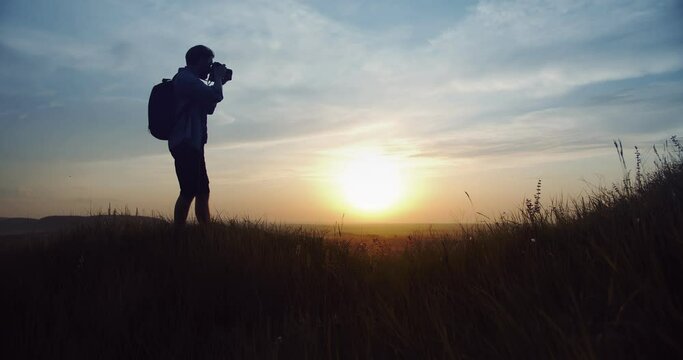 Silhouette of male photographer in summer outfit standing on high hill and taking pictures of amazing sunset. Concept of photography and adventure.