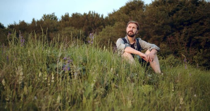 Relaxed bearded man in casual clothing sitting among green grass and enjoying beautiful nature around. Mature photographer resting on hill during summer sunset.