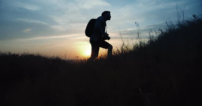 Side view of mature photographer in silhouette walking on green hill with backpack and digital camera. Bearded man in casual outfit making pictures of summer sunset.