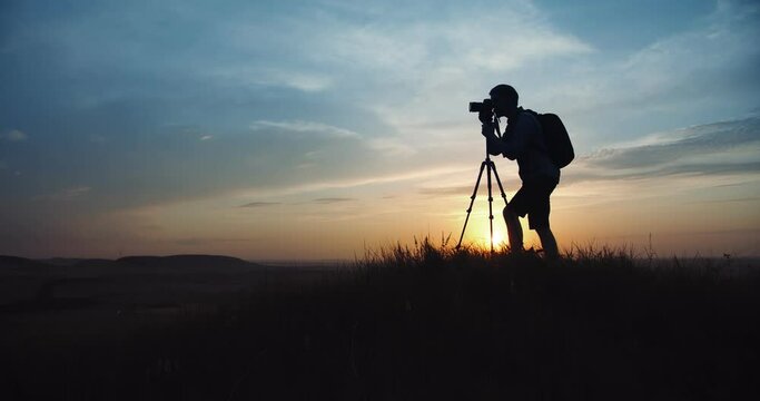 Competent photographer standing on high hill and using tripod and digital camera for shooting sunset. Concept of working process and favorite hobby.
