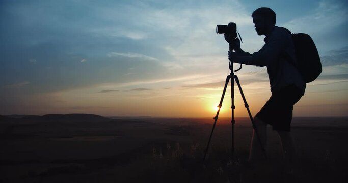 Silhouette of mature man in casual outfit with big backpack taking pictures of amazing sunset while standing on high hill. Photographer using professional tripod and digital camera outdoors.