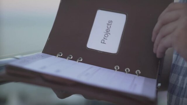 Close up shot of man hand flicking through daily planner journal notebook diary pages in warm evening sunset at seascape