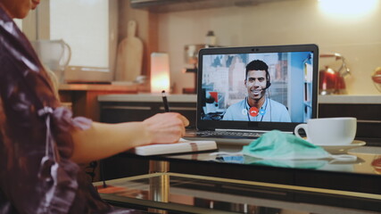 Close-up shot of Video call on computer with mixed-race colleague at home on quarantine. Woman's hands making notes. Protective mask on the table.