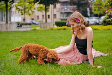 A girl in a chic long pink dress is sitting in the yard with her dog, a brown poodle. The girl walks with the dog, the hostess and the animal. A girl is playing with her dog.