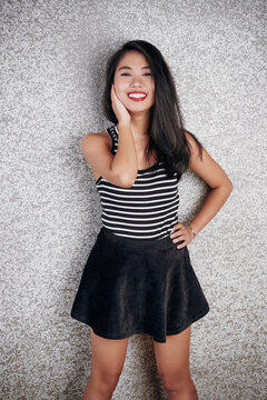 Joyful Asian girl wearing striped top and black short skirt standing against gray wall background smiling on camera