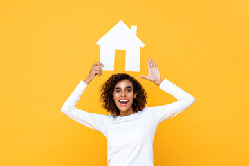 Smiling young woman holding house cutout model overhead on isolated yellow background - Powered by Adobe