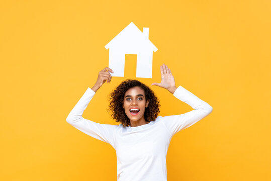 Smiling Young Woman Holding House Cutout Model Overhead On Isolated Yellow Background