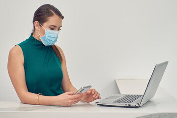 Mujer ejecutiva revisando y contestando mensajes en el celular, con mascarilla debido a la...